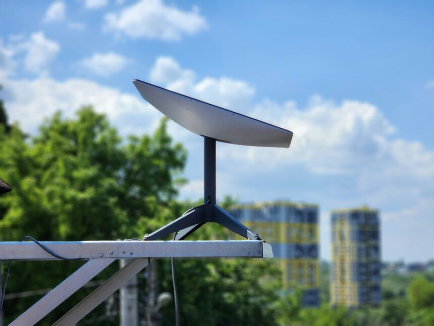 Flat satellite internet dish mounted on a pole against a blue sky with clouds and green trees