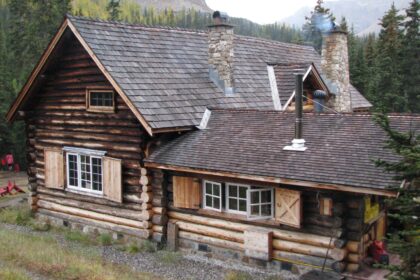 Log lodge with a steep shingled roof, two stone chimneys and shuttered windows, with forest and rock slopes behind