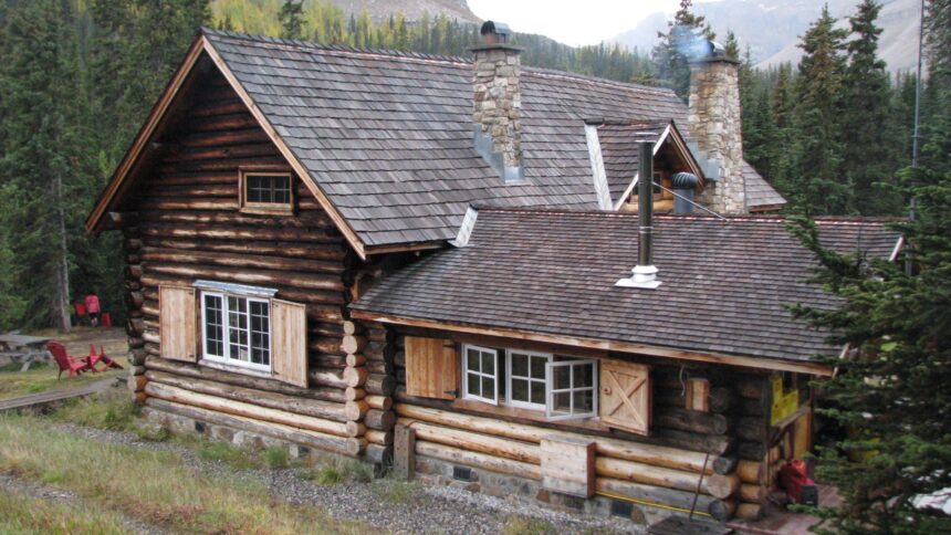 Log lodge with a steep shingled roof, two stone chimneys and shuttered windows, with forest and rock slopes behind
