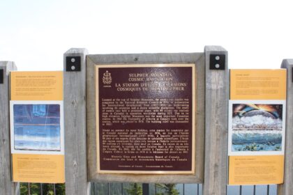 Dark bilingual commemorative plaque for the Sulphur Mountain Cosmic Ray Station on a wooden frame between two orange interpretive panels, with mountains and forest behind