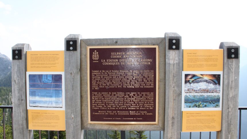 Dark bilingual commemorative plaque for the Sulphur Mountain Cosmic Ray Station on a wooden frame between two orange interpretive panels, with mountains and forest behind