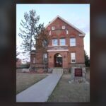 Red brick two-and-a-half-storey Territorial Court House in Fort Macleod with a central porch, a front walk and a stone marker, and a Town of Fort Macleod sign on the wall