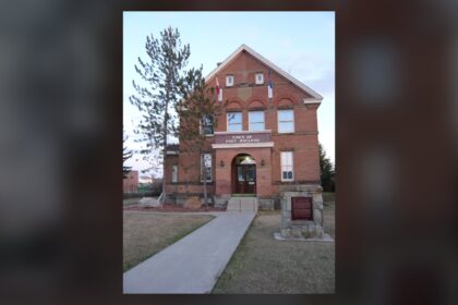 Red brick two-and-a-half-storey Territorial Court House in Fort Macleod with a central porch, a front walk and a stone marker, and a Town of Fort Macleod sign on the wall