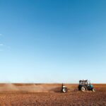 Tractor pulling a tillage implement across a dry field as dust blows behind it under a wide blue sky