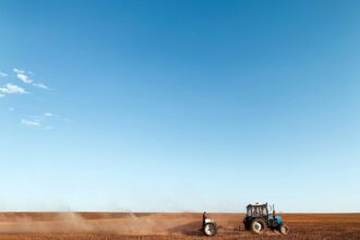 Tractor pulling a tillage implement across a dry field as dust blows behind it under a wide blue sky
