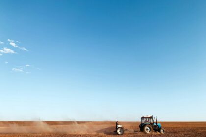 Tractor pulling a tillage implement across a dry field as dust blows behind it under a wide blue sky