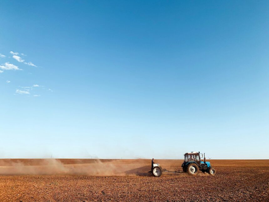 Tractor pulling a tillage implement across a dry field as dust blows behind it under a wide blue sky
