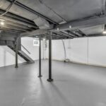 Unfinished basement with a painted concrete floor, steel support posts, exposed joists and a staircase