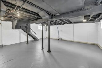 Unfinished basement with a painted concrete floor, steel support posts, exposed joists and a staircase