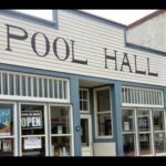 White false front with the words POOL HALL in dark capitals above a row of windows, with an OPEN sign in one pane and a red brick building at right