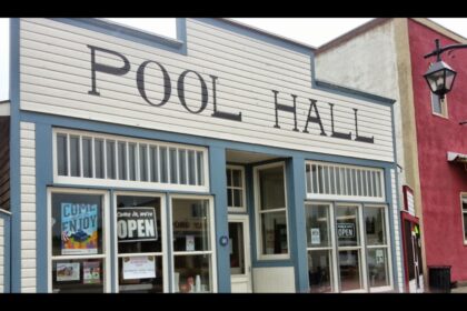 White false front with the words POOL HALL in dark capitals above a row of windows, with an OPEN sign in one pane and a red brick building at right