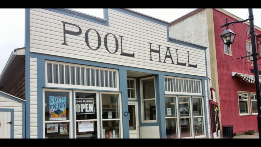 White false front with the words POOL HALL in dark capitals above a row of windows, with an OPEN sign in one pane and a red brick building at right