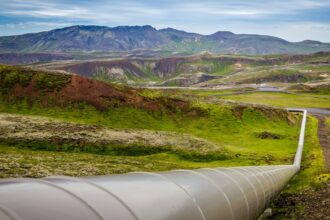 Large steel pipeline running across a green hillside landscape under a partly cloudy sky